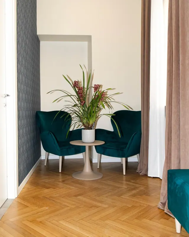 Two teal velvet armchairs flank a round side table holding a potted plant with red flowers, in a corner with herringbone wood flooring and beige curtains.