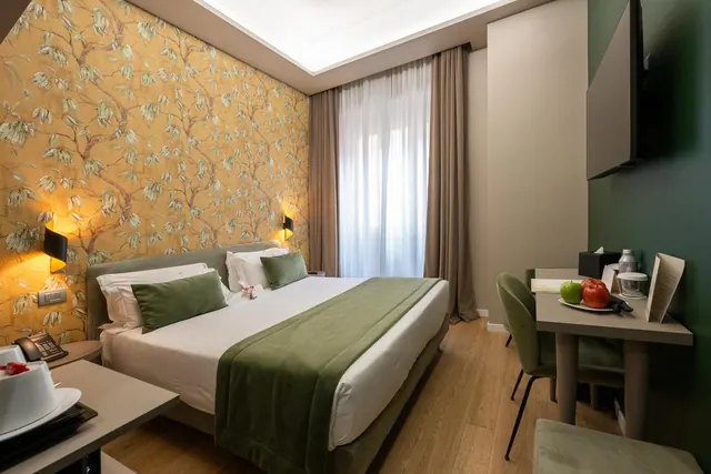 A double bed with a green throw and cushions sits against a gold floral wallpaper wall, with a wall-mounted television and a desk with fruit on the right.