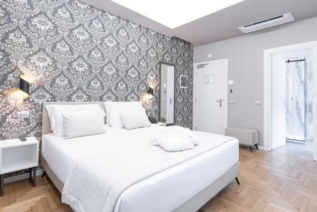 A bedroom with a made-up king bed with white linens and folded towels, damask wallpaper on one wall, and a marble walk-in shower visible through an open doorway.