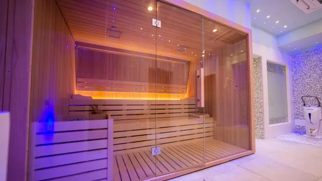 A wooden sauna with glass walls and warm lighting on its benches stands beside a mosaic-tiled wall in a spa area.