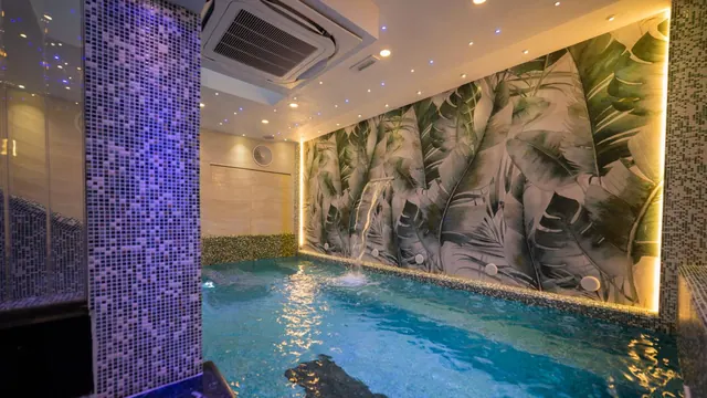 An indoor pool with a leaf-patterned wall mural, a water jet, mosaic-tiled walls and a ceiling air conditioning unit.