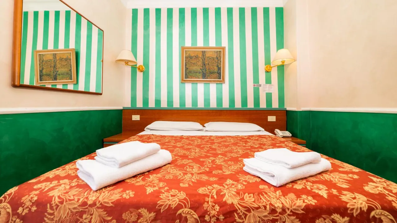 A double bed with an orange floral cover and two folded white towels sits in a room with green striped walls, framed paintings and bedside lamps.