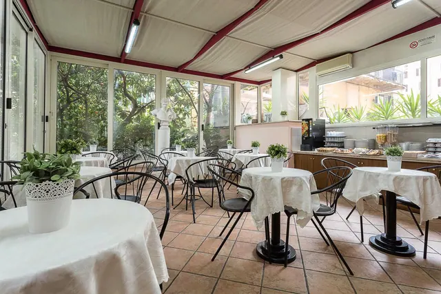 A breakfast dining room with white-clothed tables, black metal chairs, terracotta floor tiles and a buffet counter on the right, with large windows looking onto trees.