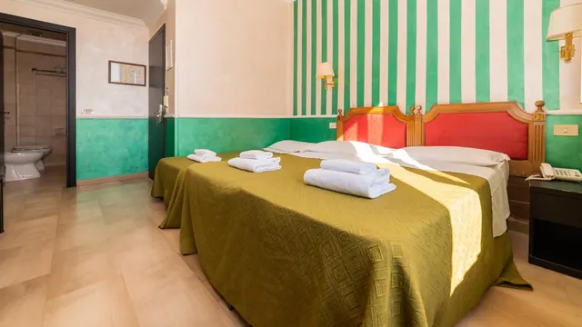 A bedroom with two beds covered in olive bedspreads and folded white towels, a green and white striped wall behind the headboard, and a bathroom with a toilet visible through the doorway on the left.
