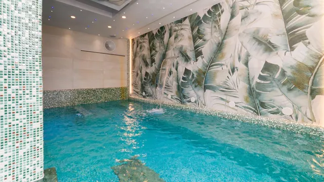 An indoor swimming pool with turquoise water, mosaic tiled walls, a small ceiling of recessed lights and a large leaf-patterned mural on the far wall.