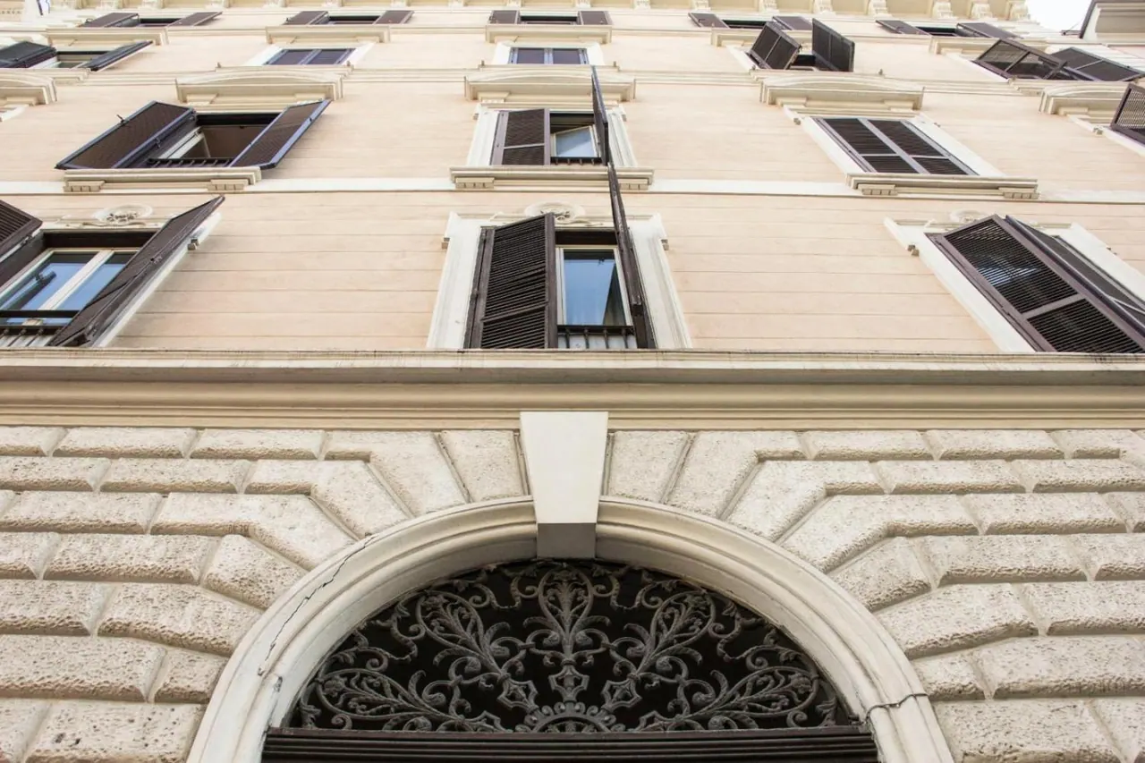Looking up at the cream-coloured facade of a multi-storey building with shuttered windows and an arched doorway fitted with an ornate iron grille.