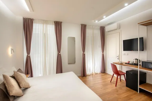 A bedroom with a double bed, sheer curtains, a wall-mounted television, a red desk chair, a kettle and a black minibar fridge.