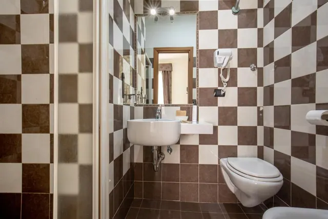 A bathroom with brown and cream checkered tiles, a wall-mounted sink and toilet, a mirror, a wall-mounted hair dryer and a frosted glass shower screen.