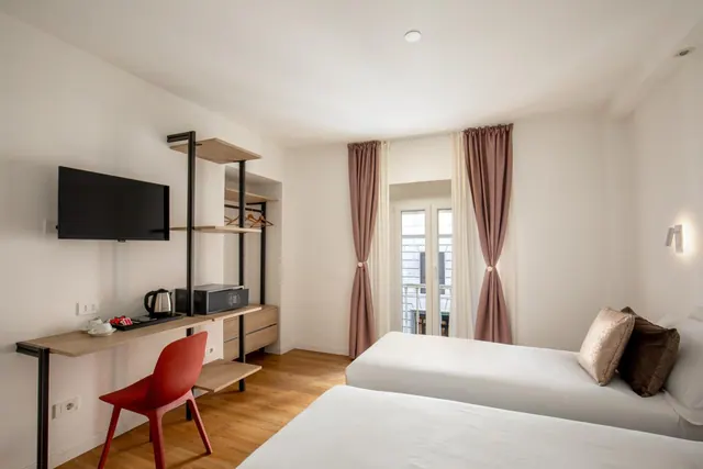 A hotel bedroom with two single beds, a wall-mounted television, a wooden desk with a red chair and a kettle, and glass doors leading to a balcony.