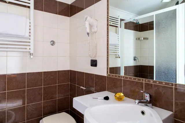 A bathroom with brown and cream tiles, a white sink with a chrome tap, a heated towel rail, a wall-mounted hair dryer, and a mirror reflecting a shower with a frosted glass door.