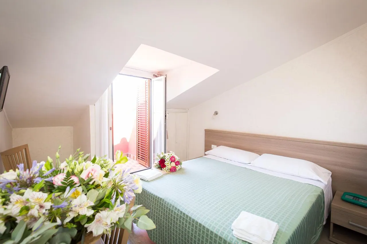 A double bed with a green bedspread and white pillows sits under a sloped ceiling, with an open balcony door and a bouquet of flowers in the foreground.