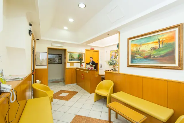 A hotel lobby with yellow armchairs and benches, a reception desk with a man standing behind it, and framed paintings on the walls.