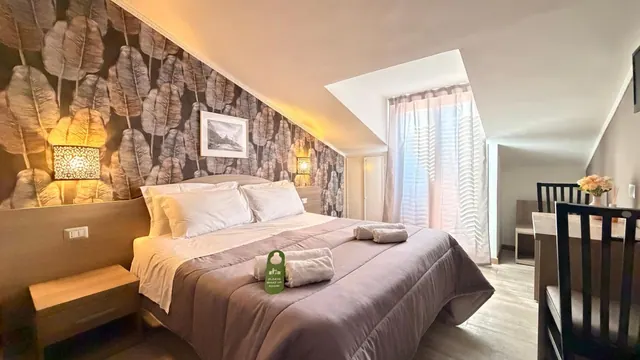 A double bed with a grey bedspread, white pillows and two folded towels sits under a sloped ceiling beside leaf-patterned wallpaper, with a desk and chair on the right.