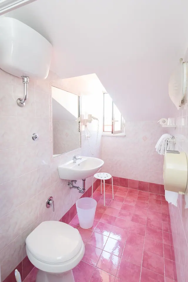 A pink-tiled bathroom with a wall-mounted sink, a toilet, a white stool, a mirror and a small window set into the sloped ceiling.