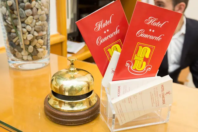 A brass desk bell, red Hotel Concorde folders and a card holder sit on a wooden reception counter beside a glass vase of pebbles, with a man in a dark suit partly visible at the right.