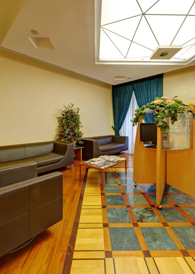 A lobby seating area with brown leather sofas and armchairs, a low coffee table with magazines, a potted plant, and a reception desk decorated with flowers under a skylight.