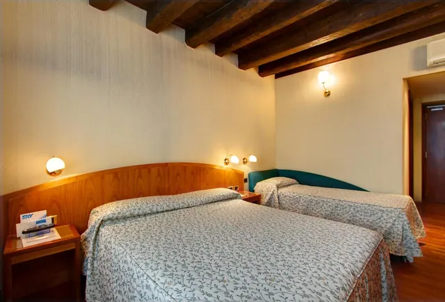 A bedroom with two beds, one with a teal headboard, under exposed wooden ceiling beams, with a wall-mounted air conditioner and a remote control on a bedside table.
