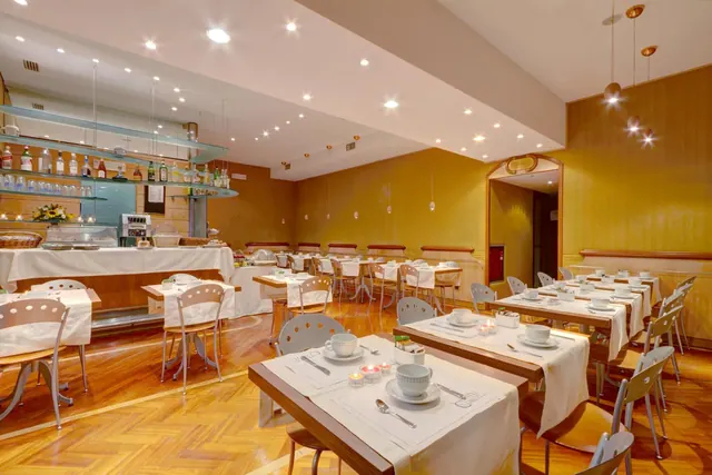 A breakfast dining room with white-clothed tables set with cups and saucers, with a buffet counter and a bar with bottles along the back wall.