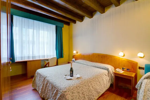 A double bed with a floral bedspread and a tray holding a champagne bottle and two glasses sits beside a wooden headboard, with a desk under the window in a room with exposed wooden beams.