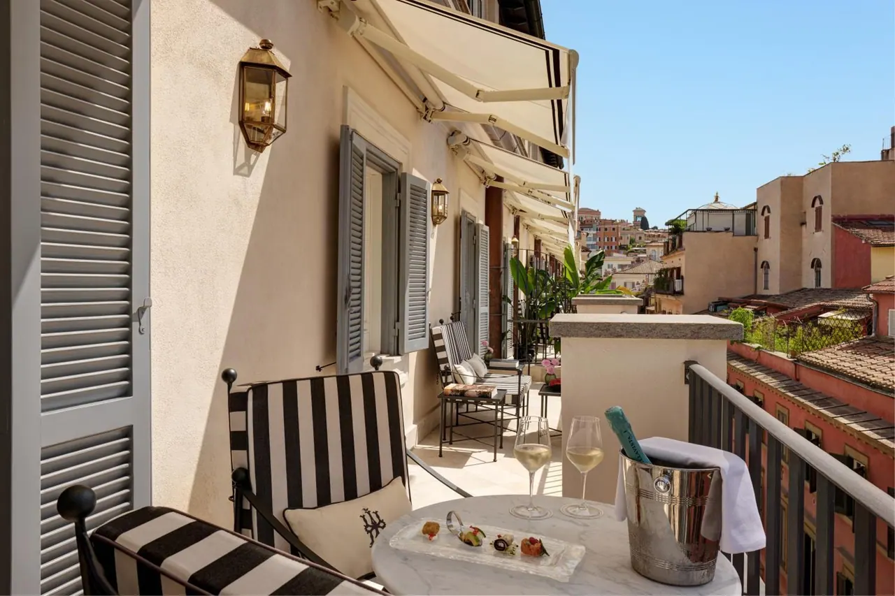 A private balcony with striped chairs, a marble table holding wine glasses and a champagne bucket, and a view over rooftops and buildings under a clear sky.