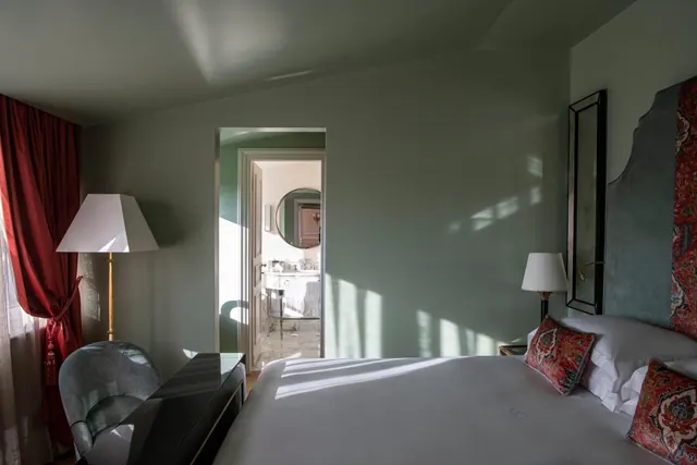 A bedroom with a made bed, patterned red cushions, a red curtain, bedside lamps and an open doorway to a marble bathroom.