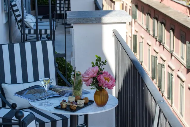 A balcony with striped lounge chairs and a round marble table holding a cocktail, a snack plate, a magazine and a vase of pink peonies, with a neighbouring pink building across the gap.
