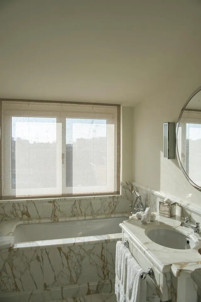 A marble-clad bathroom with a bathtub beneath a window with sheer blinds, a round mirror and a marble vanity with an inset basin.