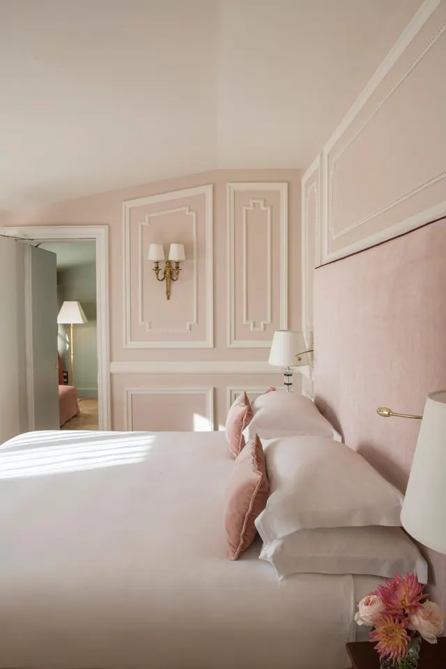 A bedroom with pink panelled walls, a made bed with white linen and pink velvet cushions, and a doorway leading to another room with a lamp and a pink chair.