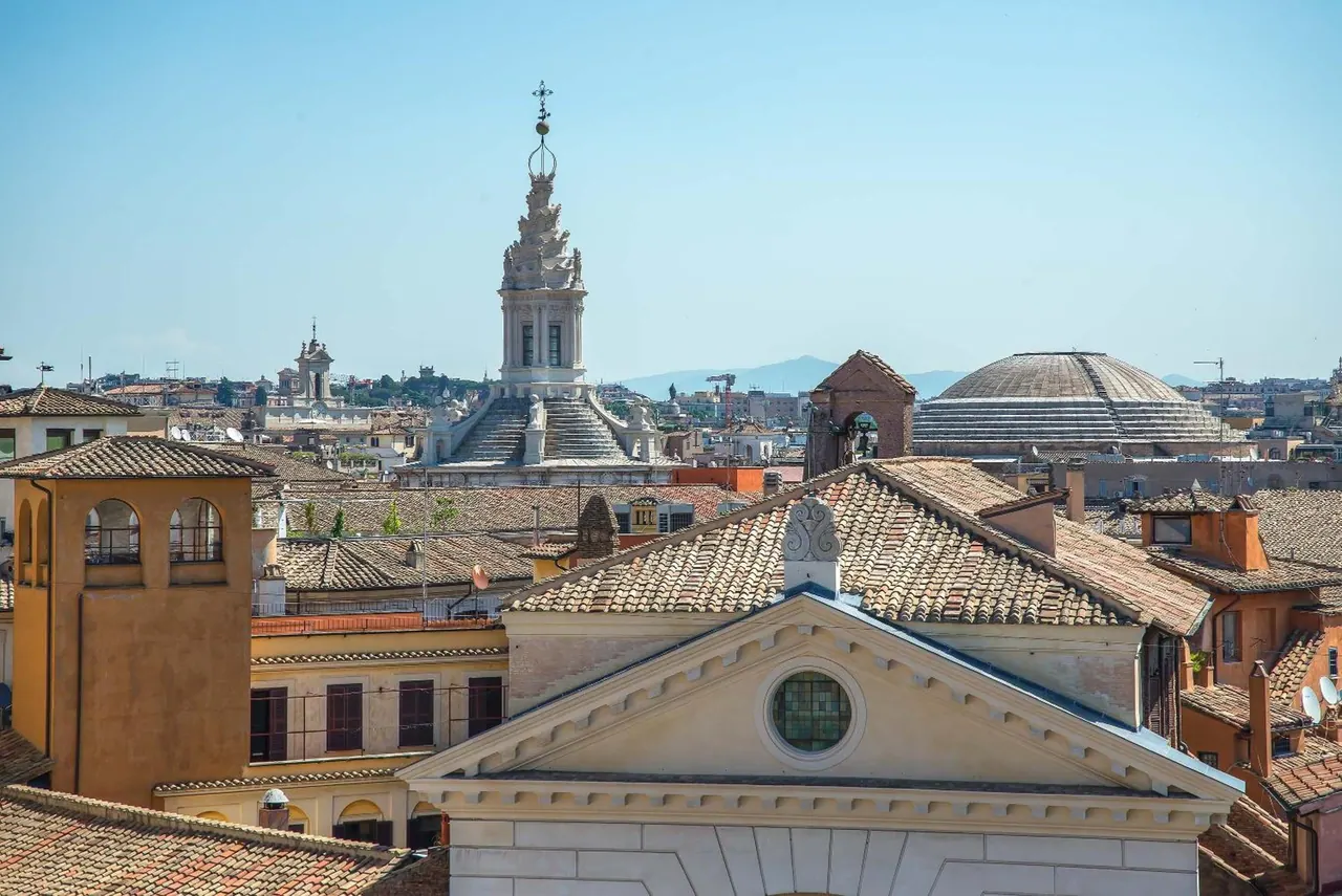 Terracotta tile roofs of Rome in the foreground, a baroque bell tower with a spire in the middle distance, and the dome of the Pantheon on the right under a clear blue sky.