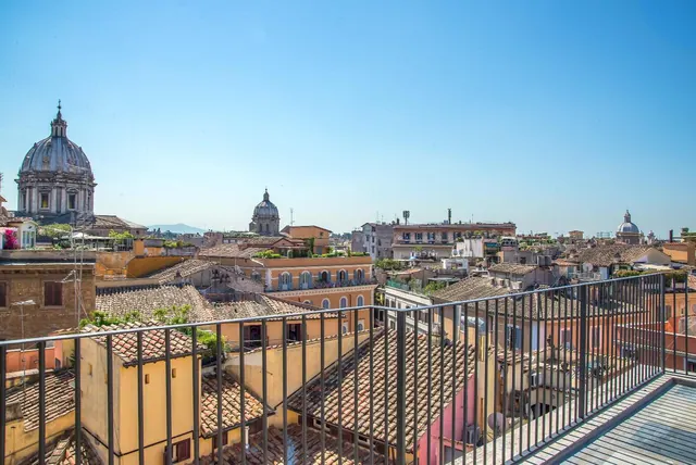 A metal railing on a terrace overlooks terracotta rooftops and several church domes under a clear blue sky.
