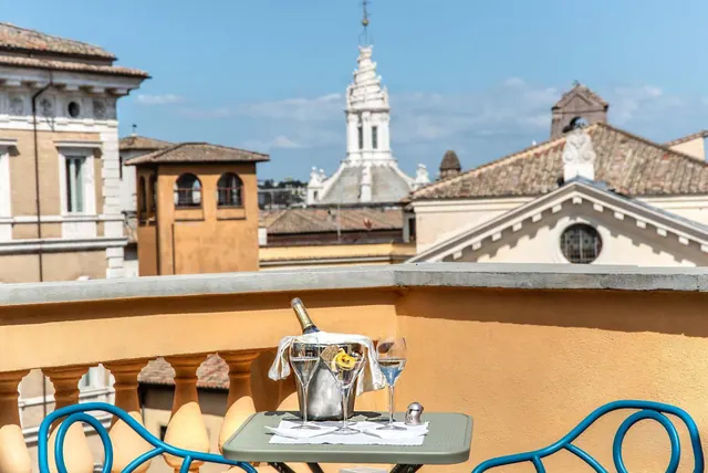 A small terrace table holds a champagne bottle in an ice bucket and three glasses, with rooftops and a church bell tower visible beyond the balustrade.