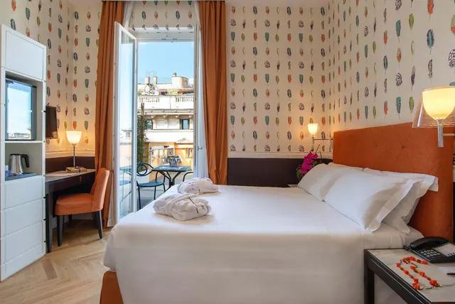 A double bed with white linens and two folded bathrobes sits beside an open door onto a small balcony with rooftop views.