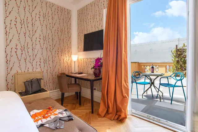 A bedroom with patterned wallpaper, a wall-mounted television, a desk with a chair, and an open glass door onto a terrace with blue chairs and a table.
