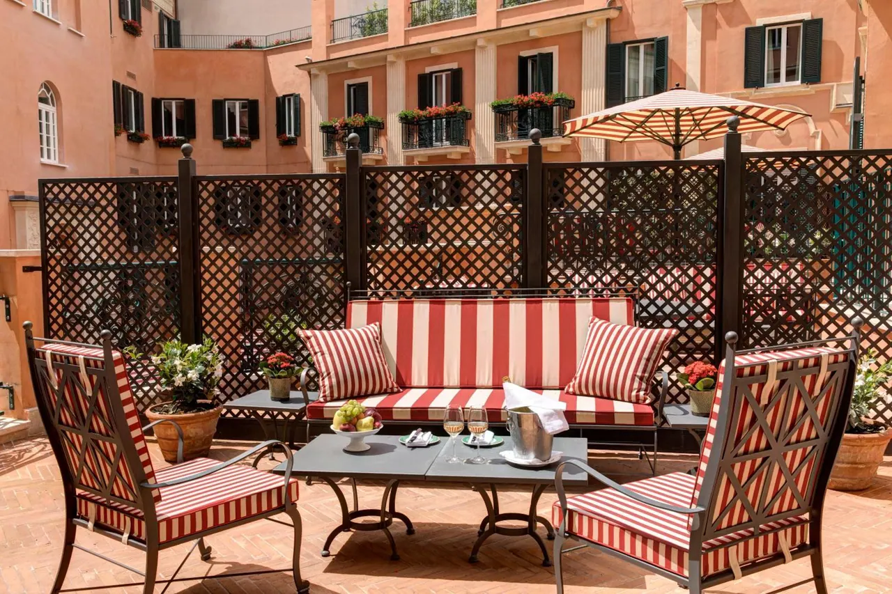 An outdoor terrace with a red-and-white striped sofa, two striped armchairs, a set table with wine glasses, an umbrella, and lattice screens facing terracotta buildings with flower-filled balconies.