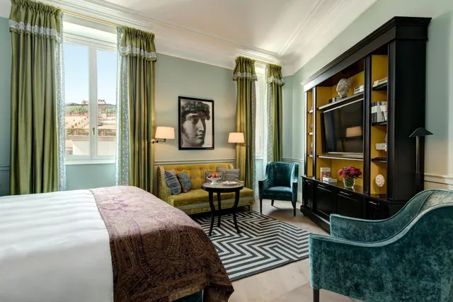 A bedroom with a large bed, a yellow sofa and round side table, teal armchairs, a dark wall unit holding a television, and a window with green curtains looking out over rooftops.