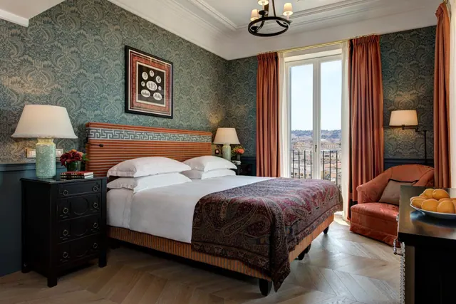 A bedroom with a made king bed, patterned wallpaper, an armchair, and French doors opening onto a balcony with a view over a city.