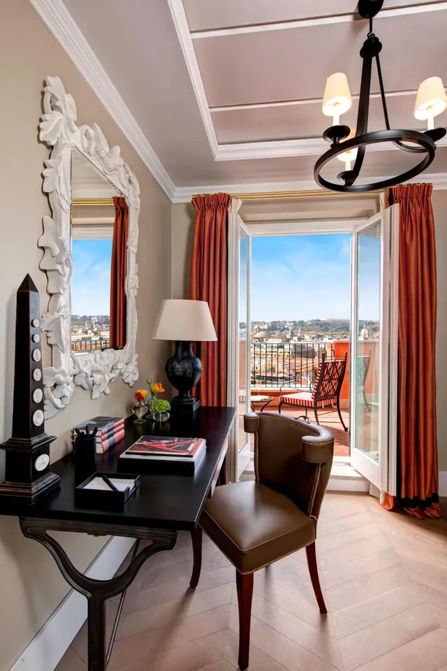 A hotel room with a dark wooden writing desk and leather chair, with open French doors onto a balcony that overlooks city rooftops.