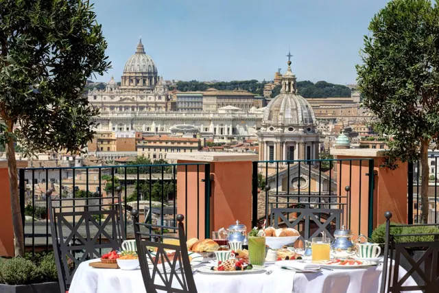 A breakfast table on a terrace looks out over St. Peter's Basilica and the surrounding Rome rooftops under a clear sky.