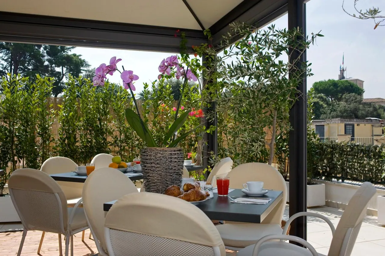 A covered outdoor terrace with cream chairs and a laid breakfast table holding croissants, orange juice and coffee, with a potted orchid, hedges and a distant bell tower behind.