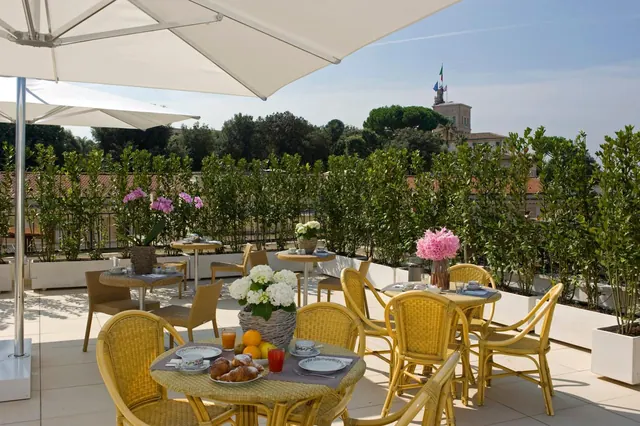 A rooftop terrace with white umbrellas and wicker chairs set around breakfast tables, with a hedge, trees and a tower with flags in the background.