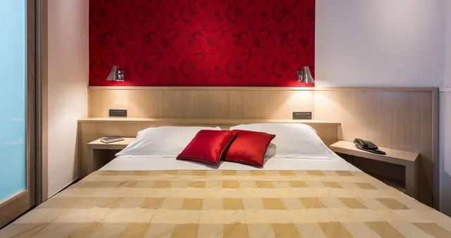 A double bed with a gold checked throw and red cushions sits against a red patterned wall with wall lamps and bedside tables on each side.
