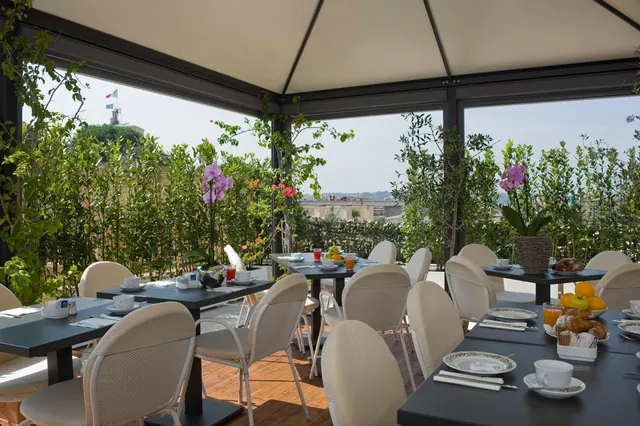 A covered rooftop terrace with black tables, cream chairs and set breakfast tables, with green plants and a view over distant rooftops.