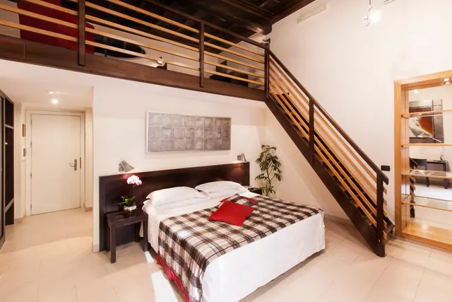 A double bed with a checked throw and red cushions sits beside a wooden staircase leading to a mezzanine level in a bedroom.