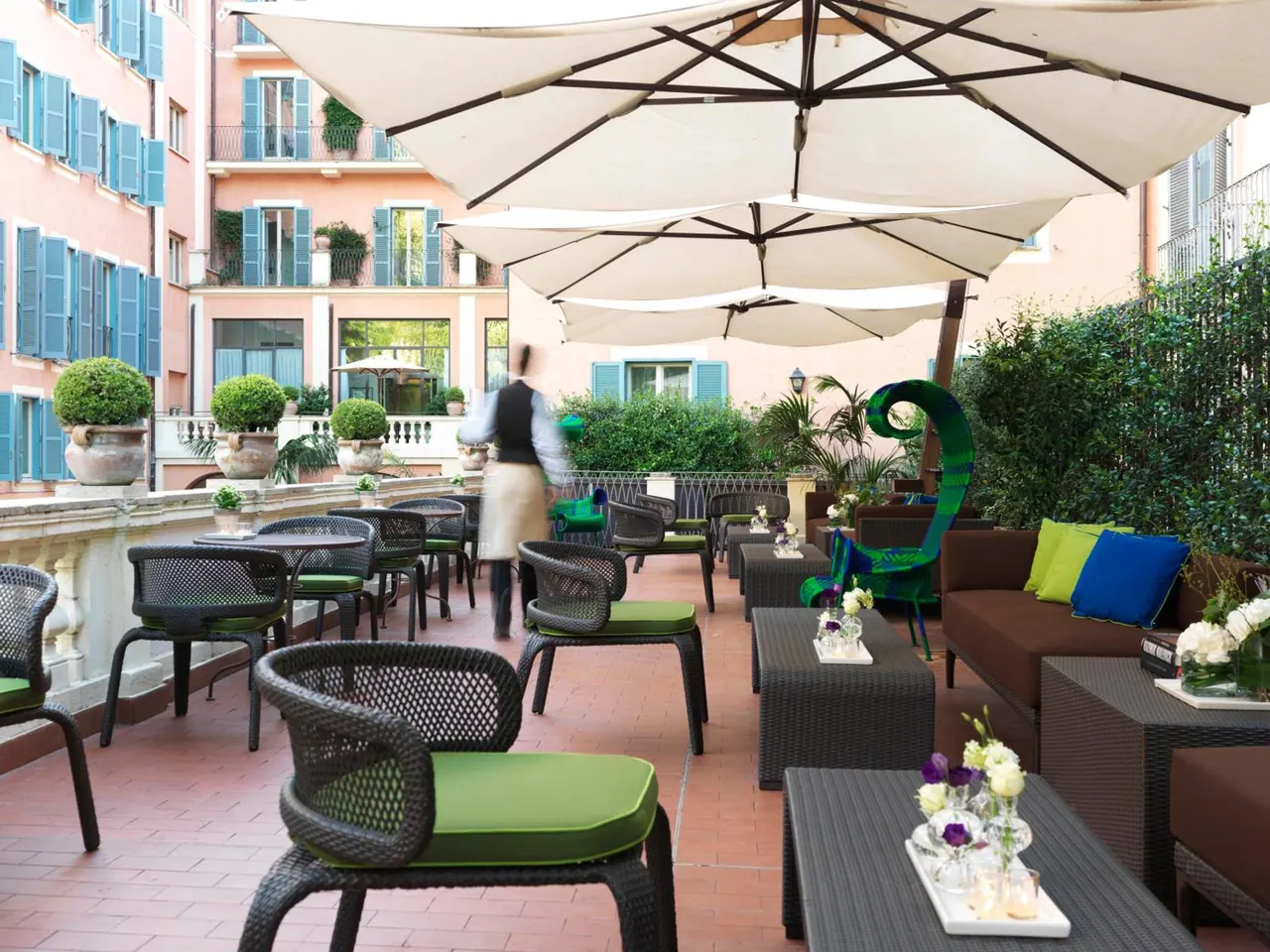 An outdoor terrace with large cream umbrellas, green-cushioned wicker chairs and lounge seating, with a waiter walking past and pink buildings with teal shutters behind.