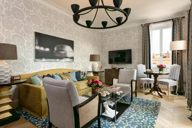 A staged living area with a yellow sofa, armchairs, a wall-mounted television and a tall window showing rooftops outside.