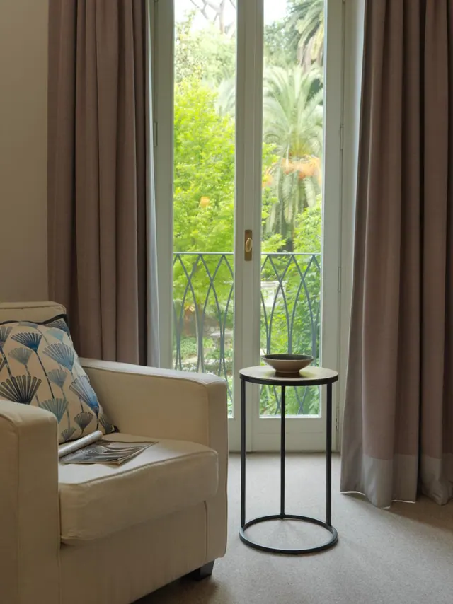 A living area with a cream armchair, a small round side table with a bowl, and French doors opening onto a balcony with a view of green trees and palms.