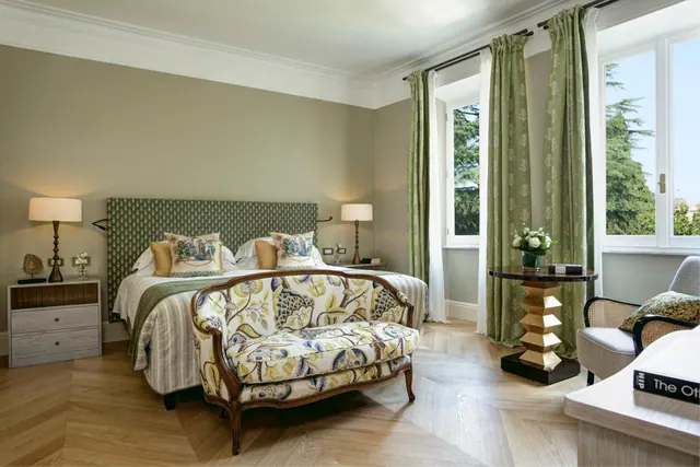 A staged bedroom with a double bed, an upholstered settee at the foot of the bed, and tall windows looking out onto trees.