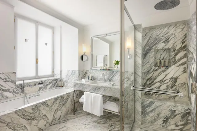 A marble-clad bathroom with a built-in bathtub, a glass-enclosed walk-in shower, a single marble vanity and a frosted window.