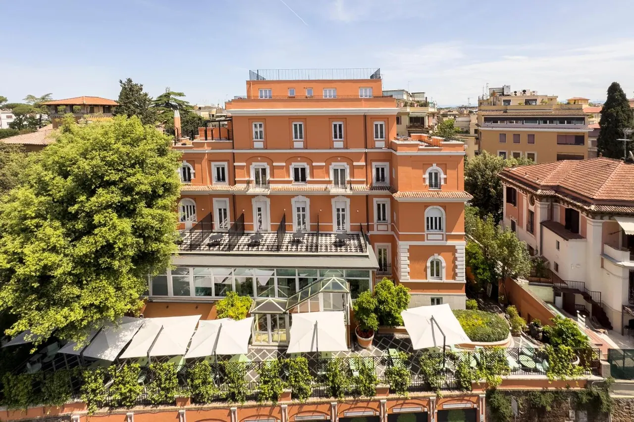 Elevated view of an orange hotel building with a terrace below set with white umbrellas and tables, surrounded by trees and neighbouring rooftops.