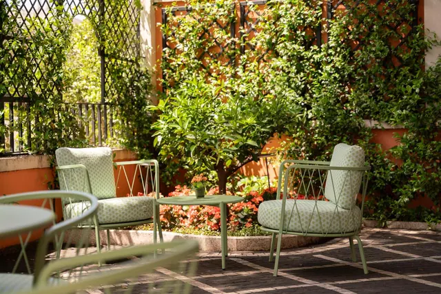 Pale green metal armchairs with cushions and a small round table stand on a paved terrace beside an orange tree, red flowers and a climbing-plant trellis against orange walls.